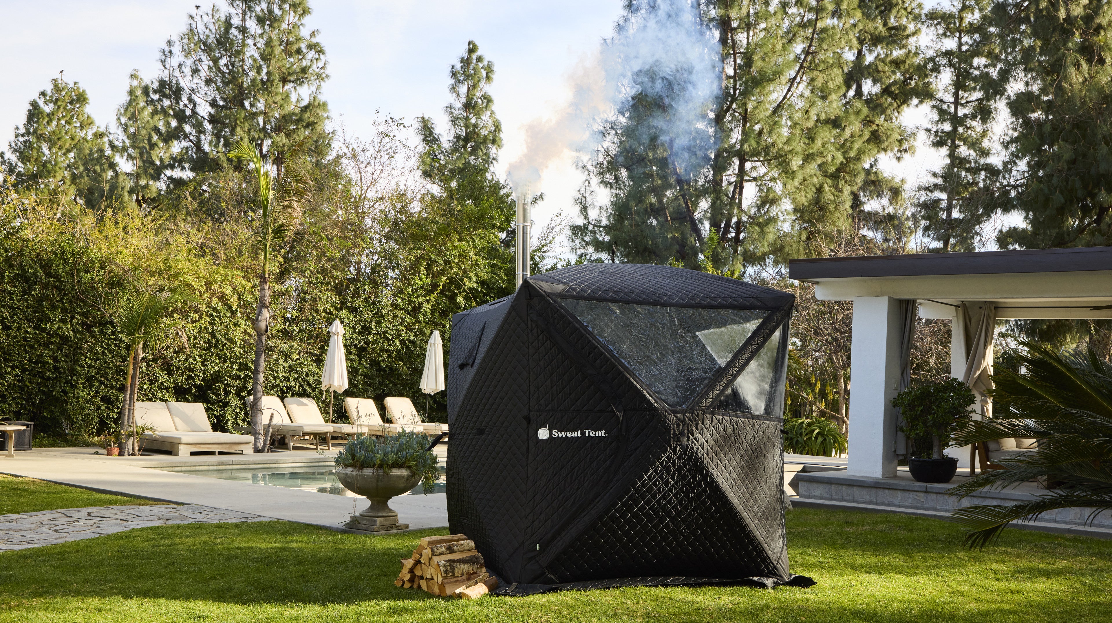 Sweat Tent portable sauna poolside on a lawn, smoke plume from the chimney - hero banner test variant (desktop)