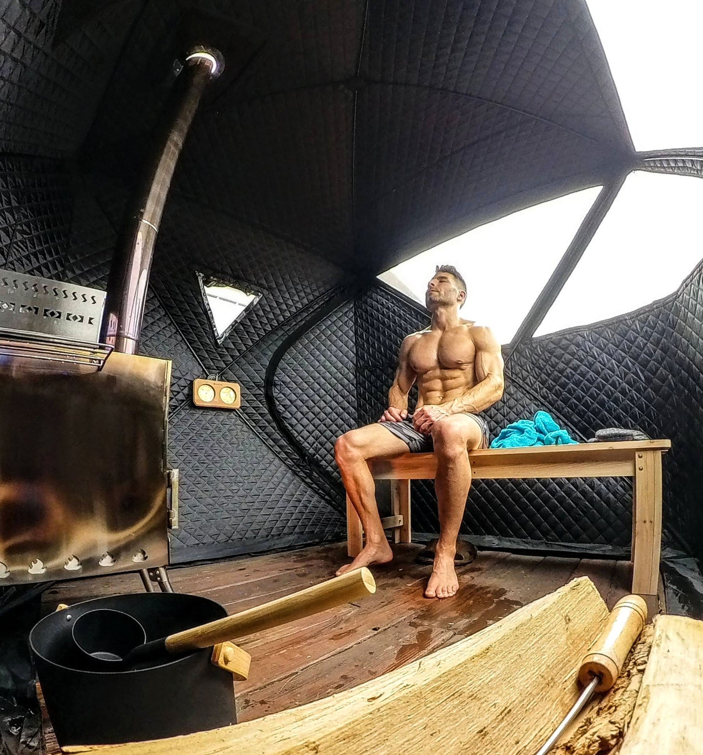 A shirtless man sitting in a Sweat Tent portable sauna with a bucket, ladle and wood stack near the fireplace